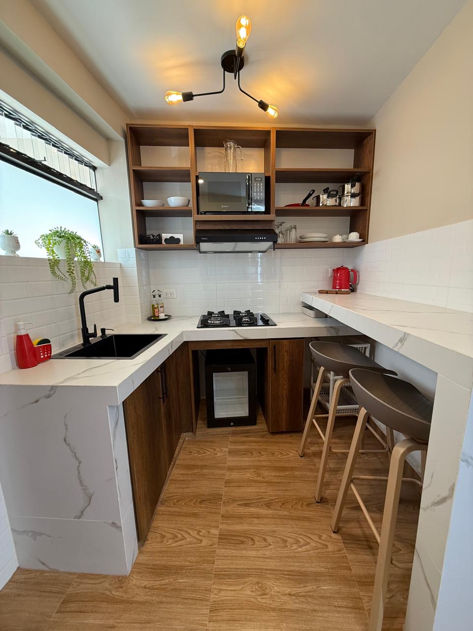 A compact kitchen features sleek wooden cabinetry and a modern countertop. Cooking appliances, including a gas stove and microwave, are neatly arranged. Open shelving displays simple dishware and a kettle. Natural light filters through a window, while stylish bar stools add seating at the countertop.
