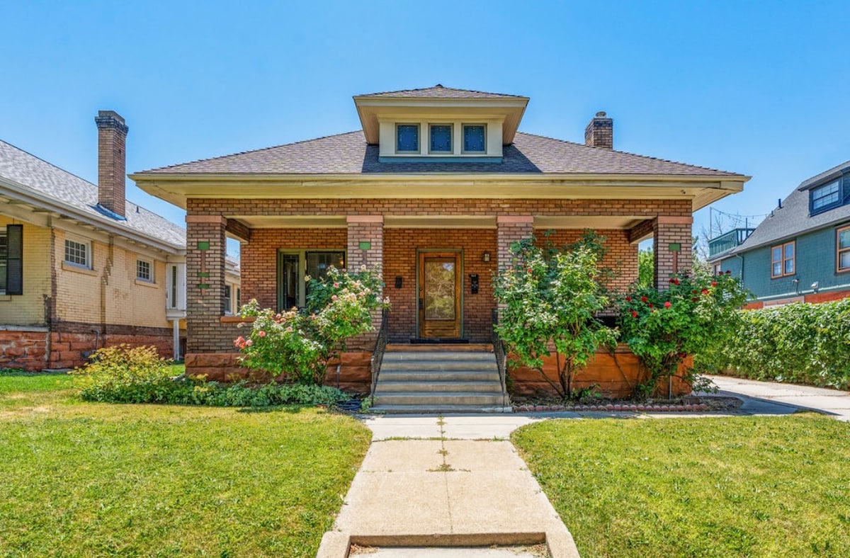 A charming craftsman-style duplex is showcased, featuring a welcoming front porch and brick exterior. Lush green landscaping and rose bushes frame the entrance. A concrete pathway leads to the main steps, while the home is surrounded by a well-maintained yard on a clear sunny day.