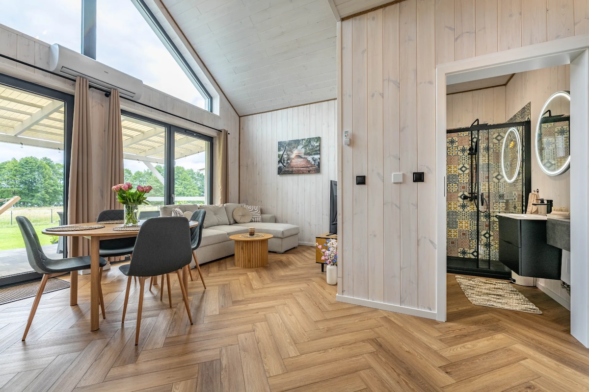 A light-filled living space features large glass doors leading to outdoor views. A dining table with four chairs sits near a cozy seating area, which includes a sofa and a wooden coffee table. A stylish bathroom area with unique tile designs is visible through an open door.