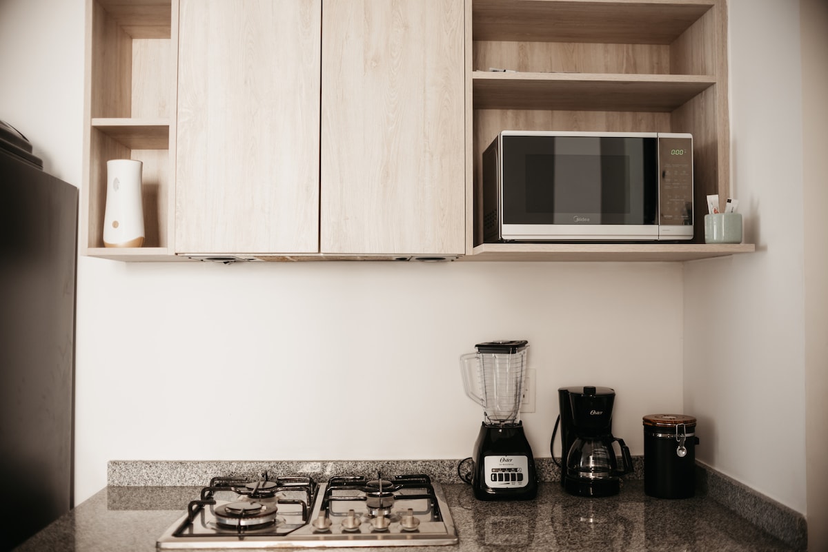 A modern kitchen features wooden cabinetry with an upper shelf displaying a microwave and small kitchen appliances. A gas stove is positioned on a granite countertop, accompanied by a coffee maker and a blender. A black container adds to the organized look.