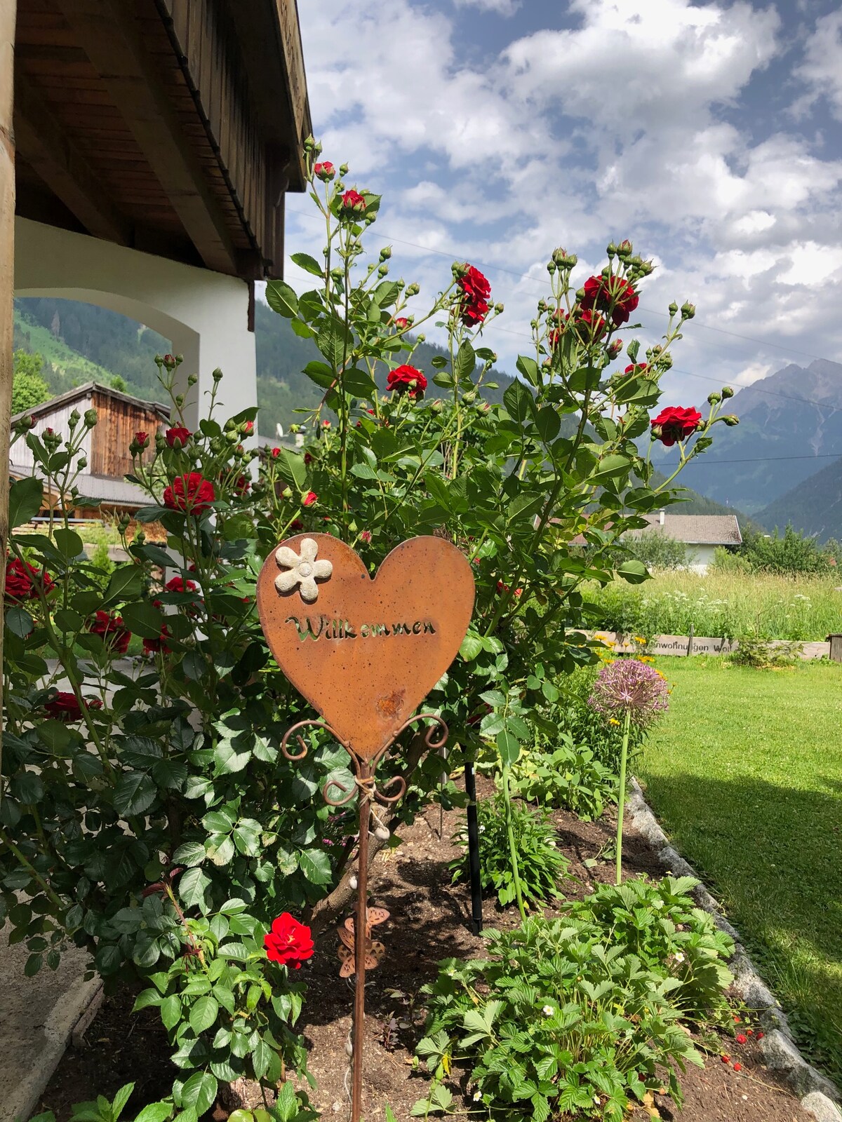 A charming heart-shaped sign is nestled among lush rose bushes, displaying the word 'Willkommen.' The garden is vibrant with green foliage and colorful blooms, set against a backdrop of rolling hills and cloudy blue skies.