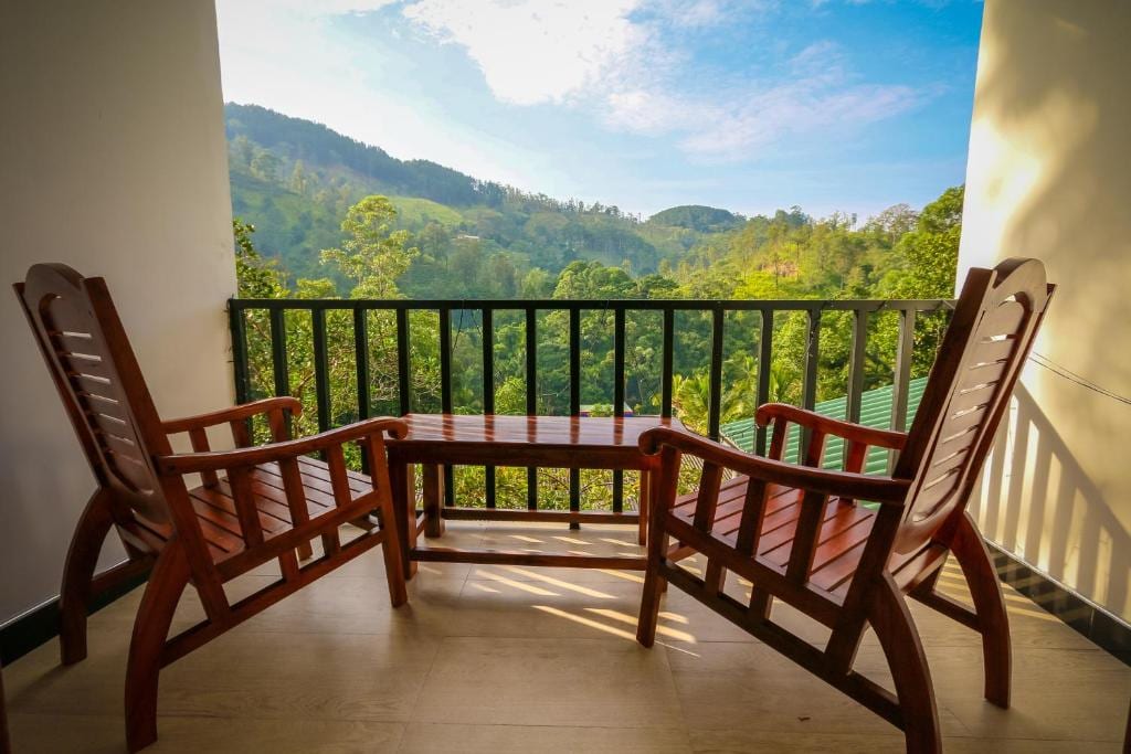 A balcony features two wooden rocking chairs positioned around a small table. The view reveals lush green hills and trees, with a blue sky and scattered clouds creating a serene backdrop.