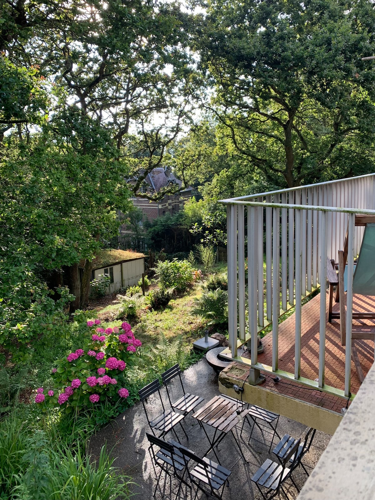 A view from the terrace showcases a spacious garden with lush greenery and vibrant pink flowers. Several black metal chairs are arranged on the patio, overlooking a serene landscape that includes trees and a small outbuilding in the background.
