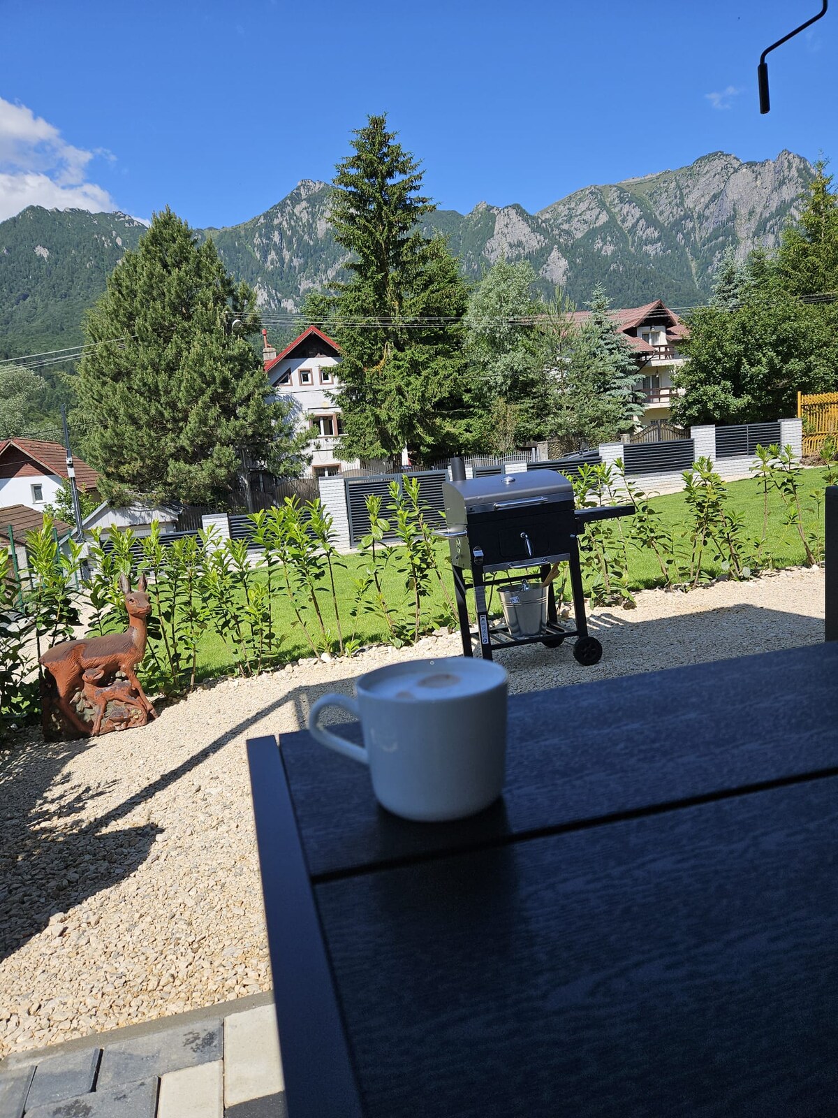 The image captures a scenic view of mountains in the background, framed by greenery. A grill stands in the yard among the landscape, and a white cup is positioned on a dark table in the foreground, inviting relaxation outdoors.