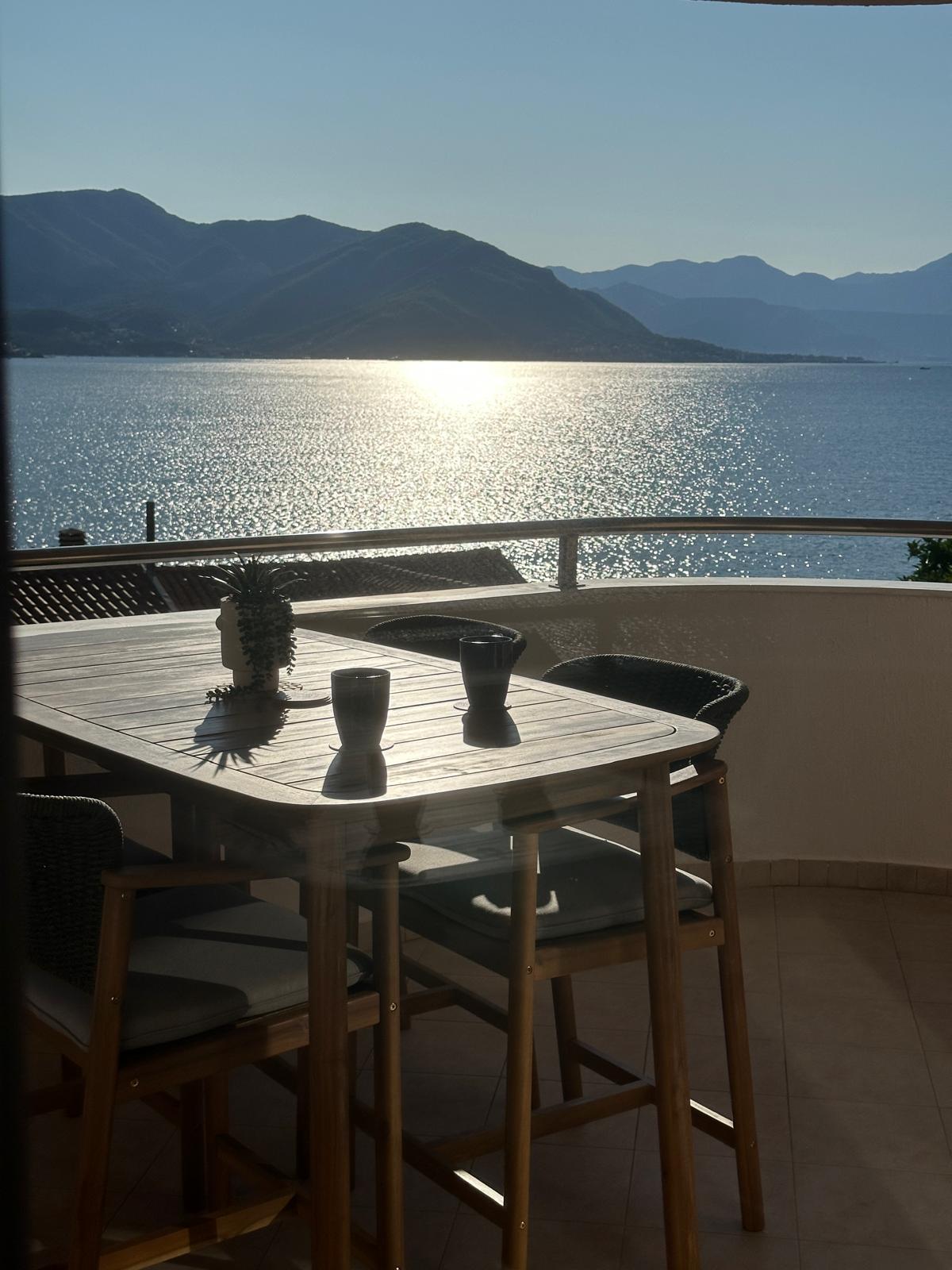 A private balcony offers a dining table with tall chairs, overlooking serene waters reflecting sunlight. The surrounding mountains create a peaceful backdrop, enhancing the coastal atmosphere. Two dark cups are placed on the table, ideal for enjoying drinks while taking in the view.