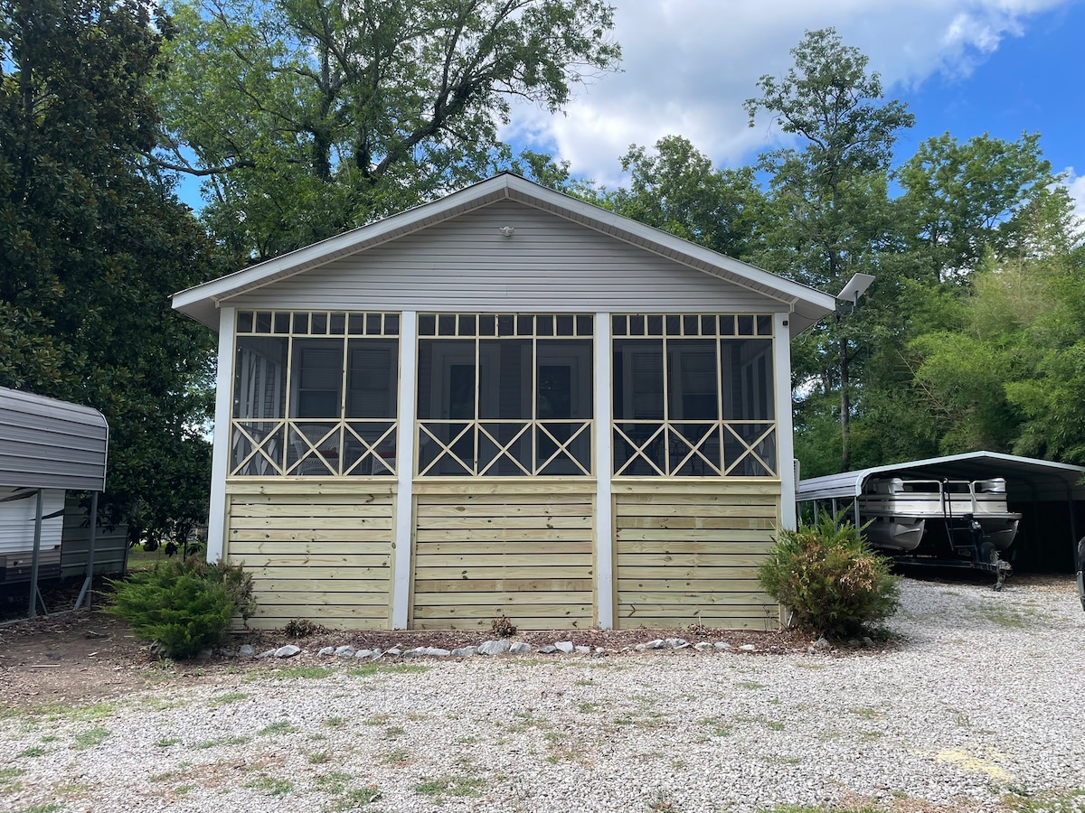 The exterior of the house features a screened porch with wooden railings and a light-colored façade. Surrounding the property are small shrubs and gravel landscaping, while boat storage is visible on the side. Tall trees provide a natural backdrop.