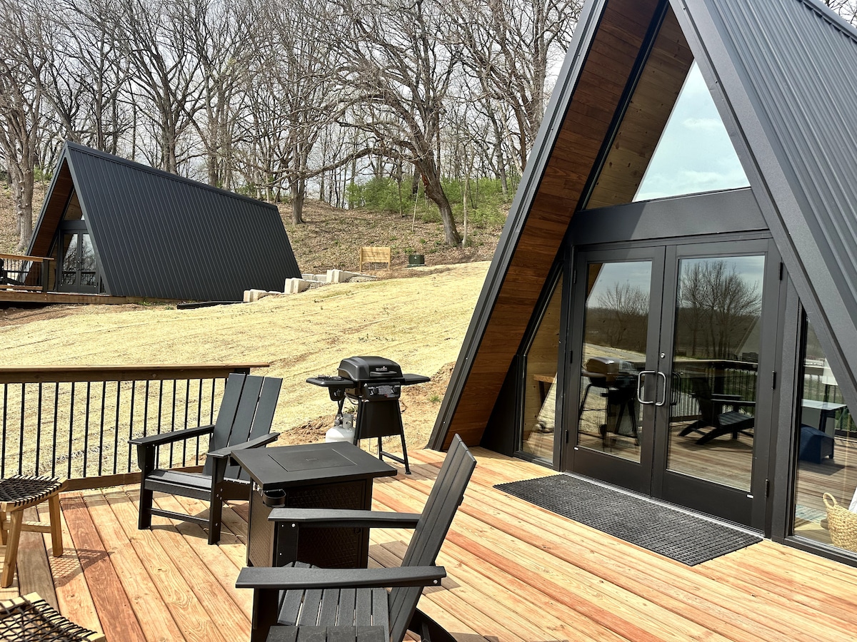 A deck area features a set of black Adirondack chairs arranged for outdoor relaxation. A grill is positioned nearby, with a backdrop of A-frame structures nestled on a gently sloping landscape. The scene is framed by a mix of trees and open space.