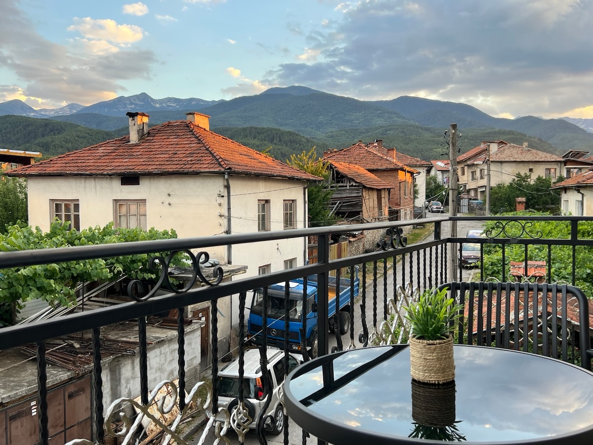 A balcony view captures the surrounding mountains under a partly cloudy sky. Traditional homes with terracotta roofs and lush greenery are visible, creating a serene atmosphere. A small potted plant sits on a round table, inviting peaceful moments outdoors.