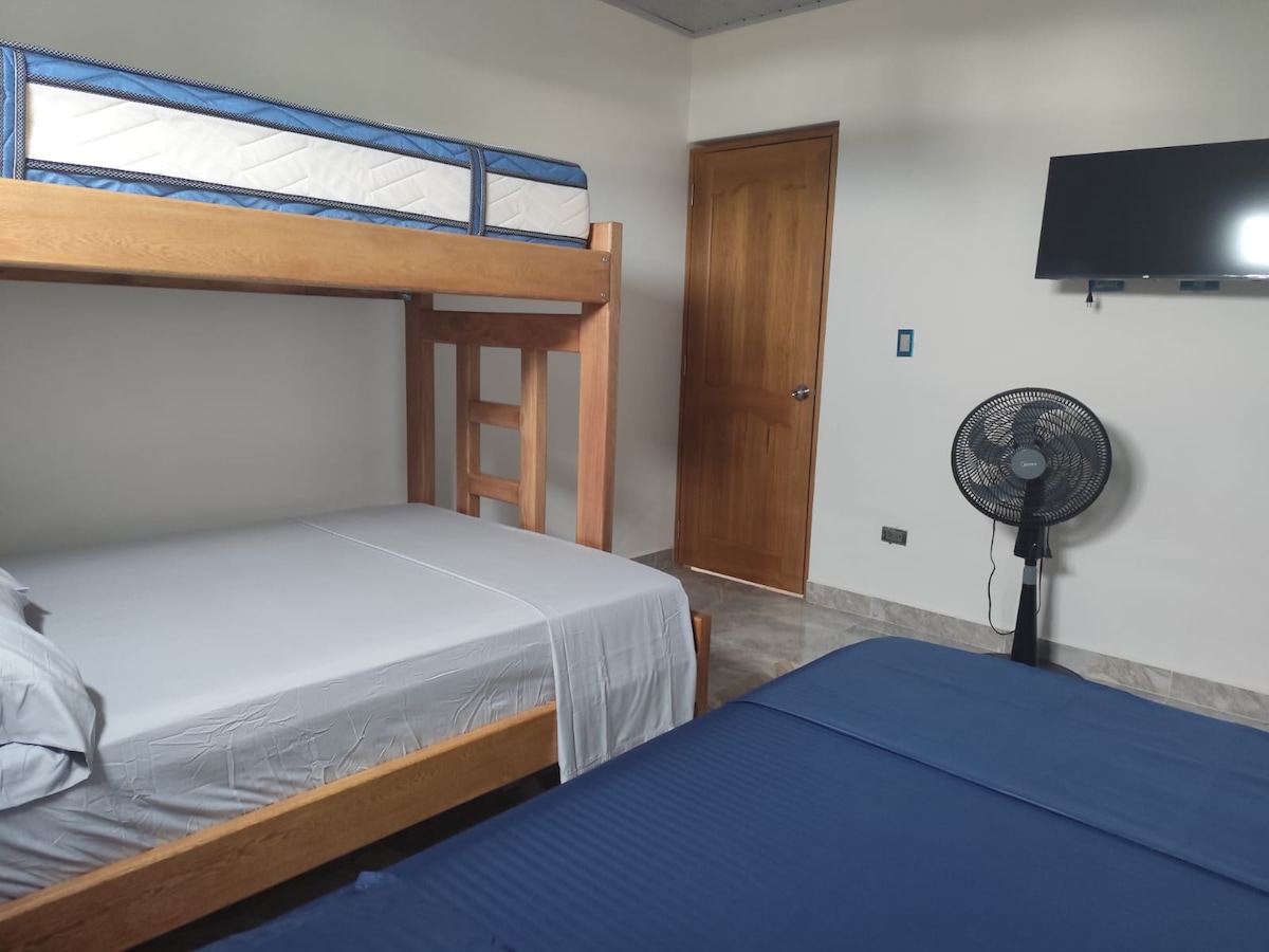 A private room showcases a double bed and a bunk bed with a double on the bottom and a single on top. A ceiling fan and a wall-mounted TV are visible, while a wooden door leads to additional areas of the property.