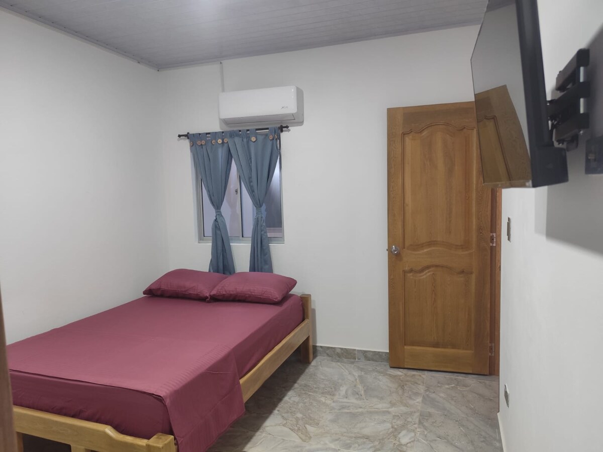 A cozy sleeping area features a wooden bed with two pillows and a rich-colored blanket. A small window is dressed with blue curtains, allowing natural light to fill the space. An air conditioning unit and a wall-mounted TV are positioned for comfort and entertainment.