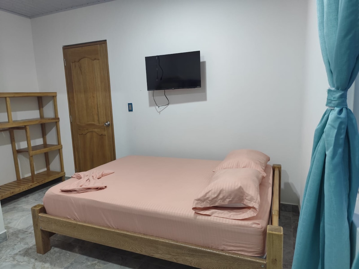 A comfortable sleeping area features a spacious bed with a light pink coverlet and two matching pillows. A wooden shelf stands against the wall, and a flat-screen TV is mounted above the bed. Light blue curtains frame the window, creating a serene ambiance.