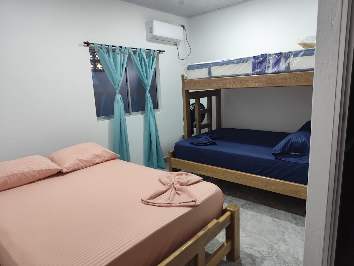 The room features a spacious double bed in a soft peach tone alongside a wooden bunk bed with a blue mattress. Light blue curtains frame the window, allowing natural light to enter. An air conditioning unit is mounted on the wall for comfort.