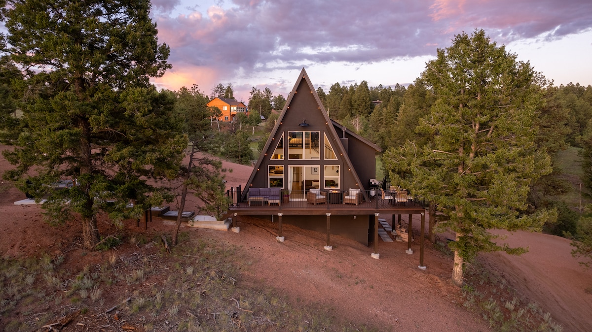 The A-frame cabin, surrounded by tall trees, features a large deck with seating overlooking the natural landscape. The triangular structure is complemented by multiple windows, inviting natural light and showcasing the tranquil mountain setting.