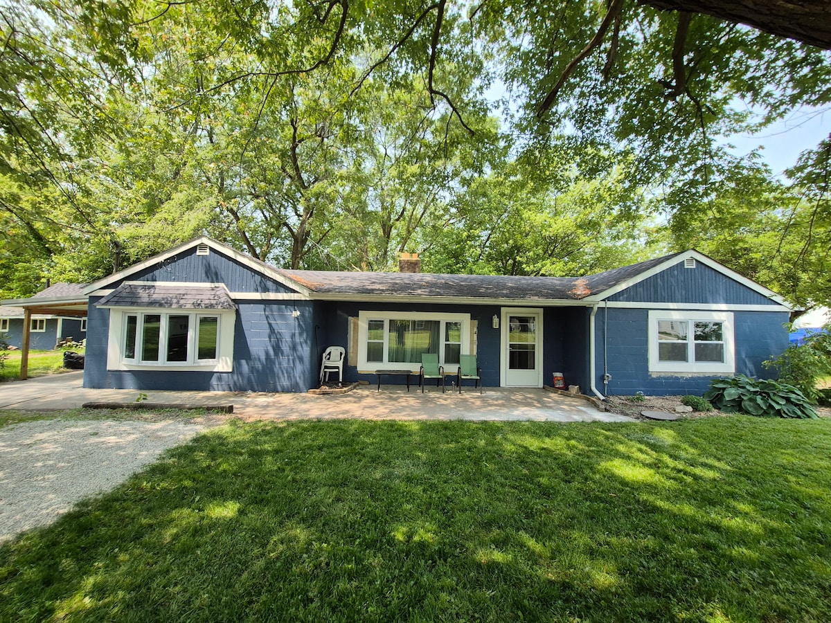 A single-story blue house is set within a landscaped yard, framed by lush green trees. The front features multiple windows and a welcoming path leading to a seating area with chairs. A spacious lawn is visible, providing a serene outdoor space.