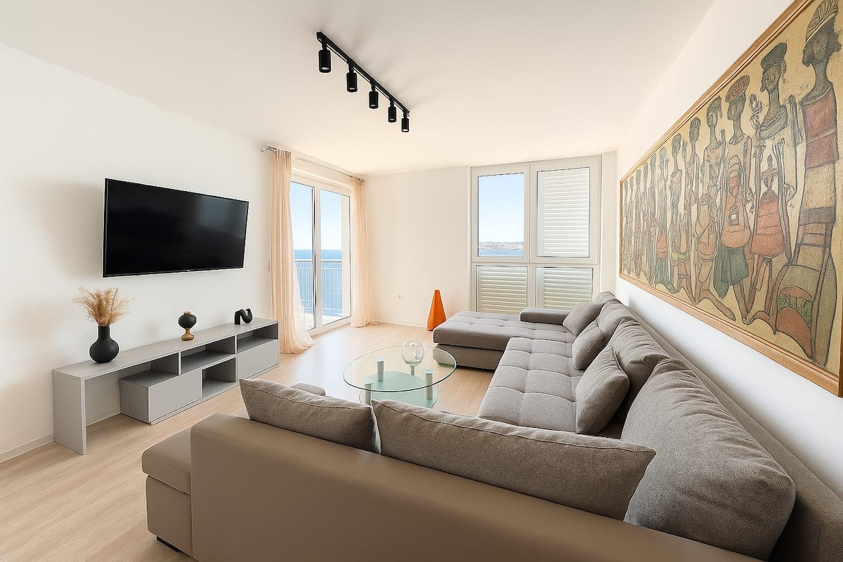 A spacious living area features a large, neutral-toned sectional sofa positioned beside a circular glass coffee table. A flat-screen television is mounted on the wall, and large windows with views of the surroundings allow ample natural light. A colorful artwork adorned with figures is displayed on one wall.