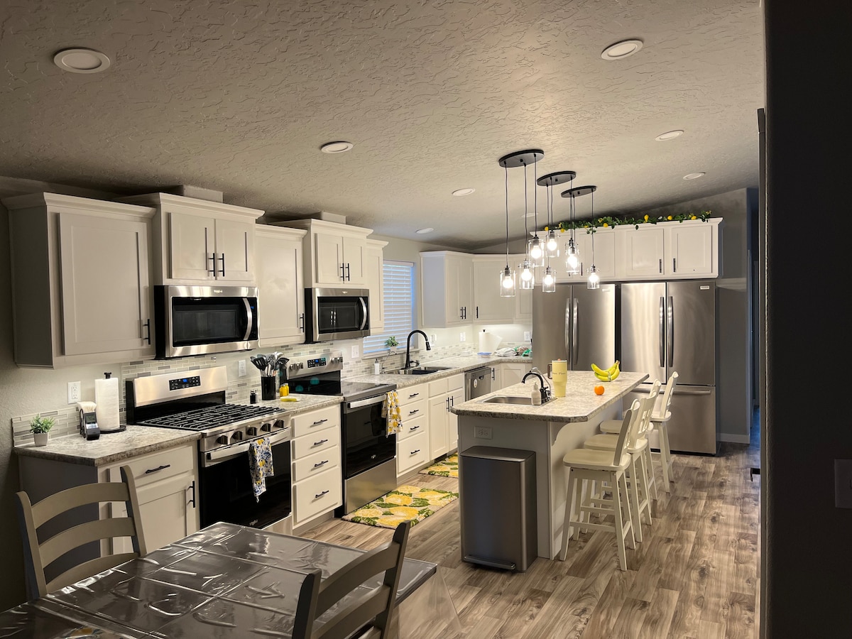 A newly designed kitchen features modern appliances, including a gas stove, double ovens, and two refrigerators. Bright, pendant lighting hangs above a central island with seating for four. A dining table is positioned nearby, surrounded by light-colored chairs, and decorative items are displayed on the countertops.