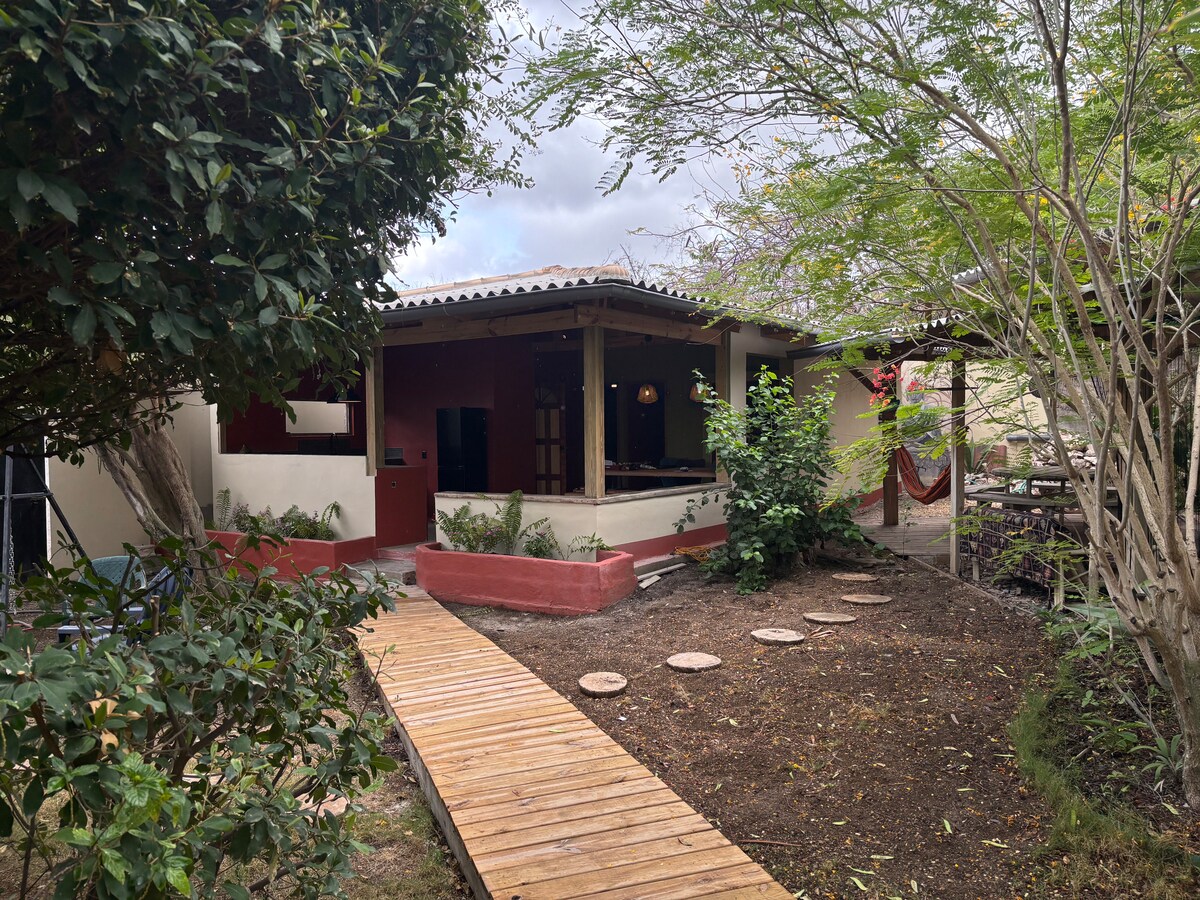 A wooden walkway leads from a garden area towards the entrance of a single-story dwelling. Lush greenery surrounds the path, with small stepping stones placed beside it. A shaded porch is visible, offering a cozy spot for relaxation.