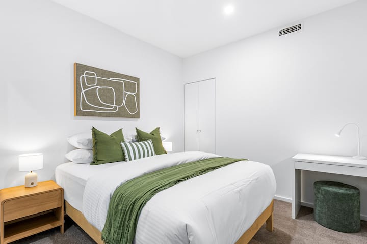 Quarto brilhante e calmante com lençóis brancos nítidos, detalhes verdes suaves e móveis simples que criam um espaço tranquilo para relaxar.