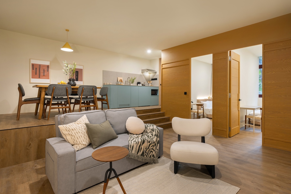 A spacious living area features a comfortable sofa, paired with accent chairs and a side table. A dining area with a large table and chairs is visible, alongside a modern kitchenette with light blue cabinetry. Two doors lead to separate bedrooms, enhancing the suite's functionality.