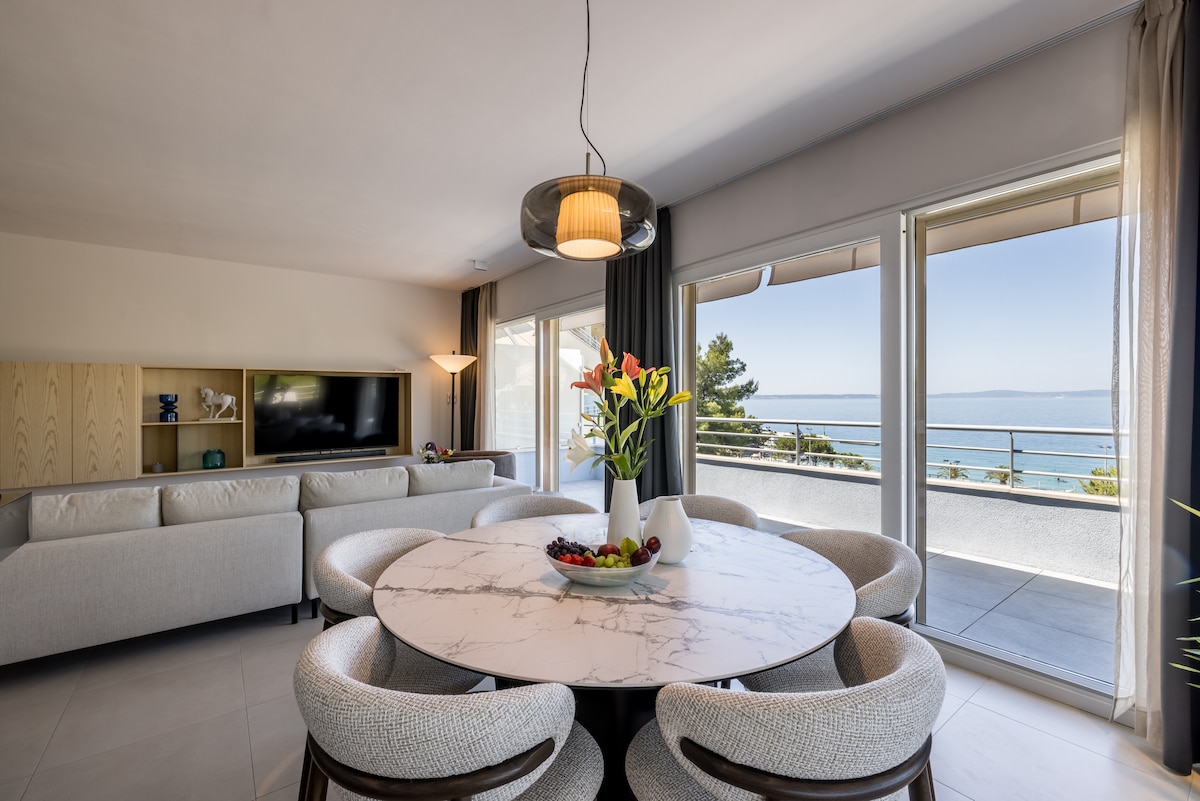 A round marble dining table with soft grey chairs is arranged centrally, complementing a light-filled living area. Large glass doors frame a serene seaside view, while a stylish light fixture hangs above the table. A cozy sofa is visible in the background, adding comfort to the space.