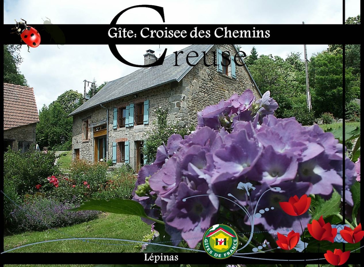 A charming gîte is depicted surrounded by lush greenery and vibrant flowers. The two-story building features blue shutters and a welcoming facade. The garden showcases a variety of blooming plants, contributing to a tranquil outdoor setting, ideal for relaxation and enjoying nature.