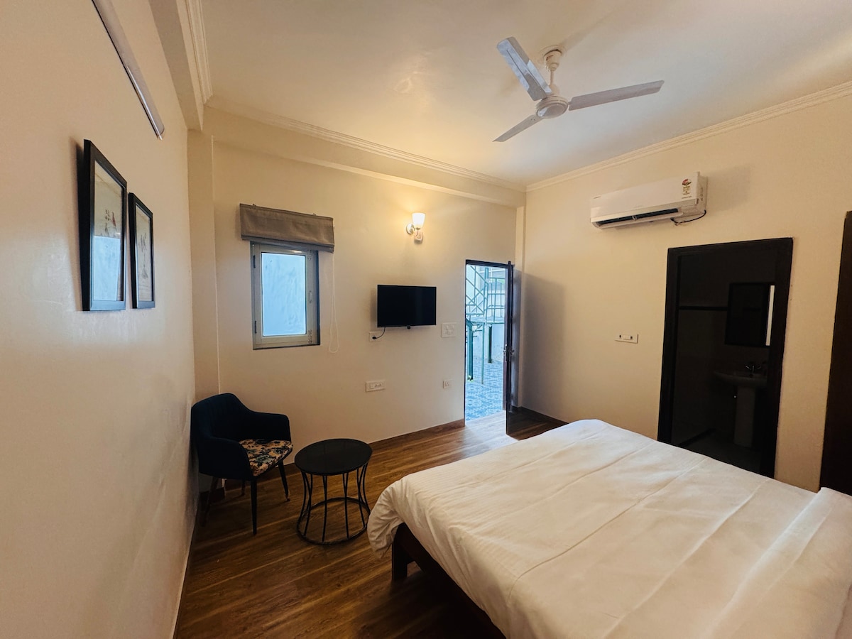 A well-lit room features a spacious bed with crisp white linens. A small table and a chair are positioned in one corner. A wall-mounted TV is visible, and a door leads to a patio. Natural light is allowed in through a window, framed by a simple blind.