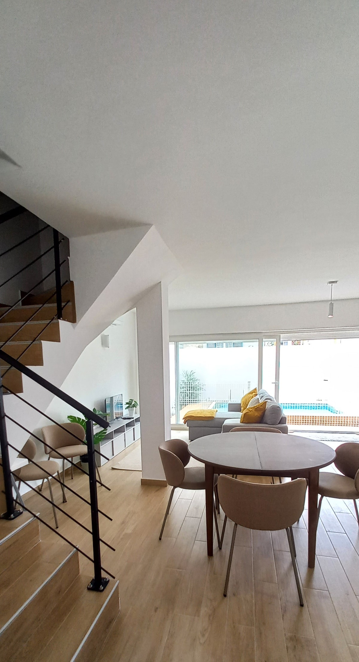 A light-filled living space is visible, featuring a round dining table surrounded by beige chairs. A staircase leads to an upper level, while large glass doors open to a pool area. A comfortable sofa with a yellow cushion adds a touch of color.
