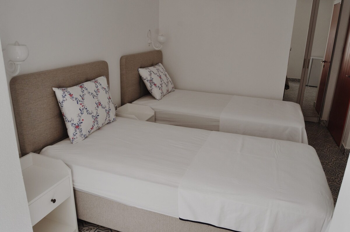 The room features two single beds with light bedding and decorative pillows. A small bedside table is positioned next to each bed. Soft lighting is provided by wall sconces, and mirrored wardrobe doors are visible in the background, enhancing the sense of space.