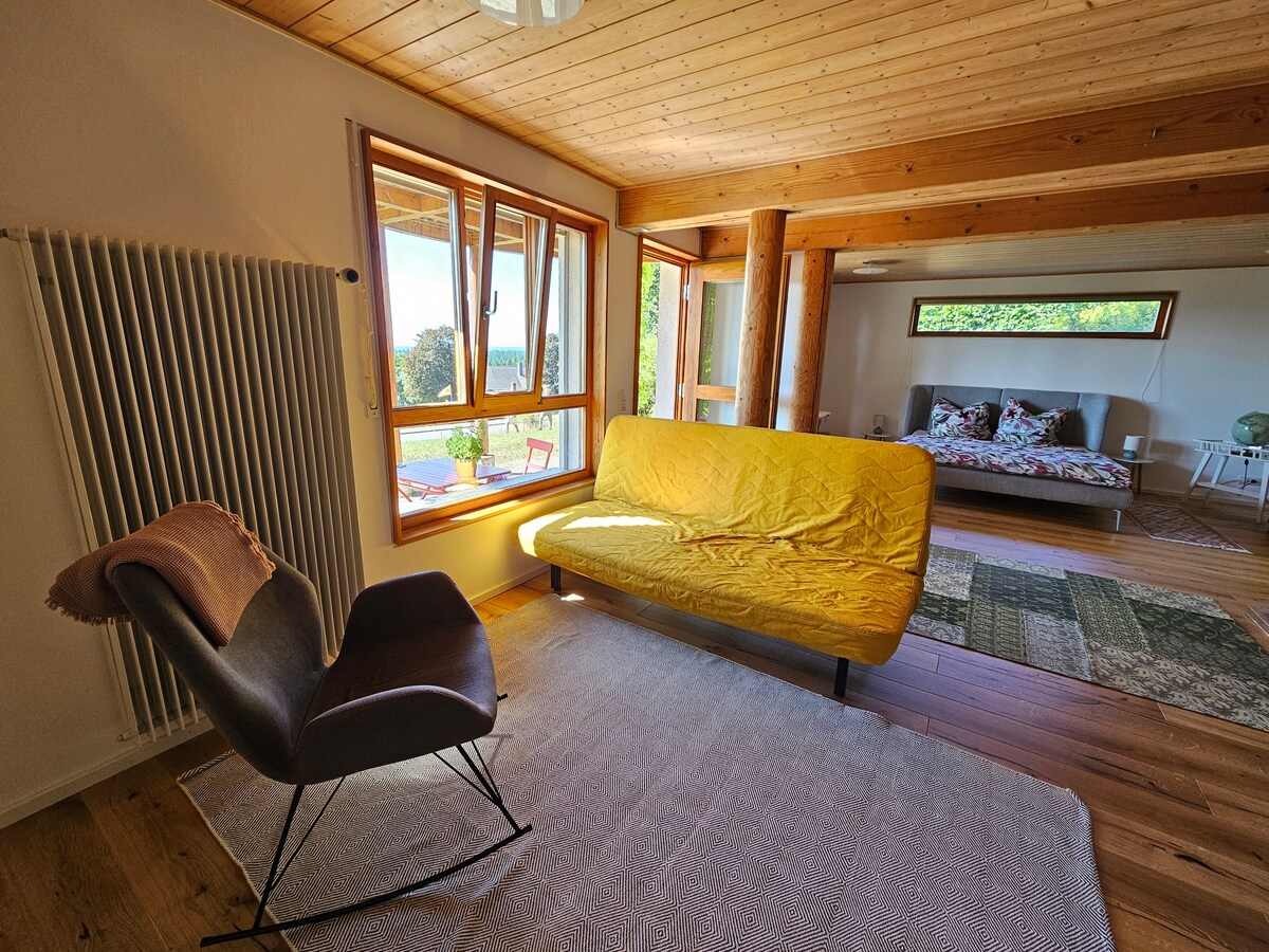 A bright living area features a yellow sofa and a black chair positioned on a light rug, with wooden floors and ceilings. Large windows offer views of the outdoors, allowing natural light to flood the space. A cozy sleeping area is visible in the background.