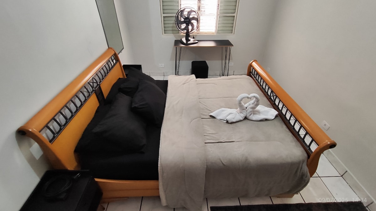 A spacious bed with a light-colored blanket and dark pillows is centrally positioned in the room. A fan is placed near a small desk against the wall, while white towels are arranged neatly at the foot of the bed. Natural light enters through a window with light-colored blinds.