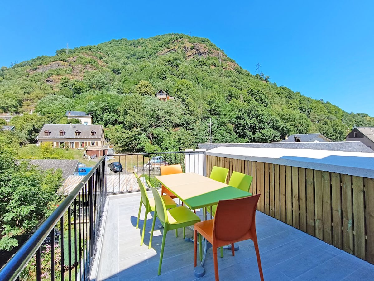 An outdoor terrace features a vibrant table surrounded by colorful chairs, providing a cheerful space for relaxation. The backdrop reveals a lush green hillside, contributing to a natural setting. The area is well-lit, showcasing clear blue skies above.