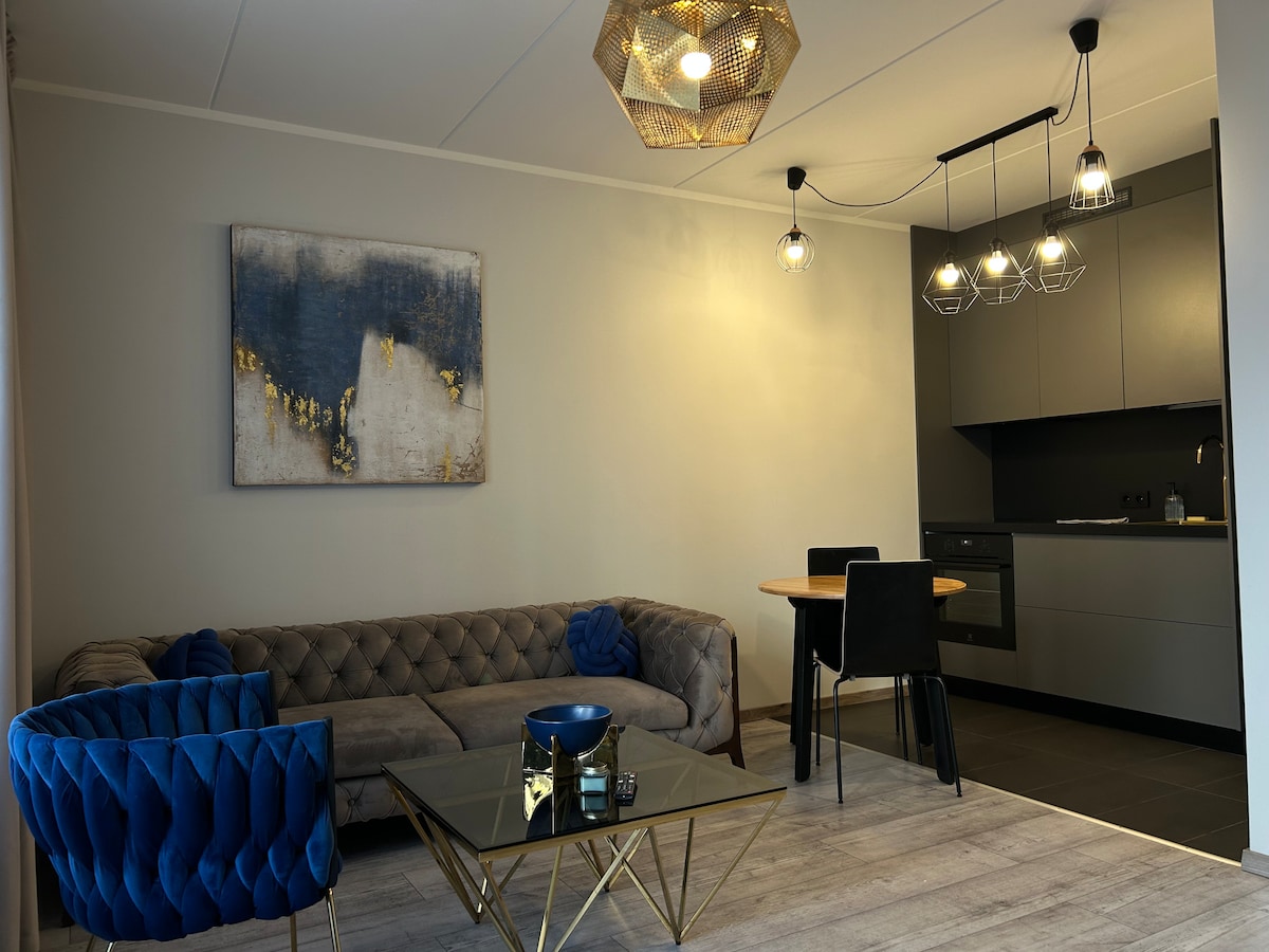 A modern living area is presented with a beige sofa, a glass coffee table, and a round dining table with black chairs. The kitchen is visible in the background, featuring dark cabinetry and modern lighting fixtures. An abstract painting is displayed on the wall.