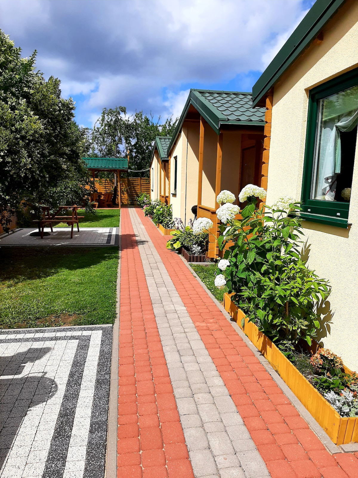 A well-maintained pathway lined with flowering plants leads to wooden cabins, surrounded by grass and landscaped areas. The inviting entrance features vibrant blooms and neatly arranged outdoor furniture, set against a backdrop of clear blue skies with scattered clouds.