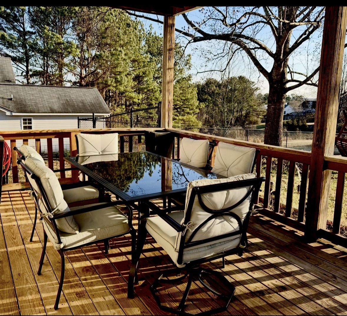 A spacious outdoor deck is furnished with a large glass-topped table surrounded by six comfortable chairs with soft cushions. Natural light filters through the trees, casting gentle shadows on the wooden floorboards.