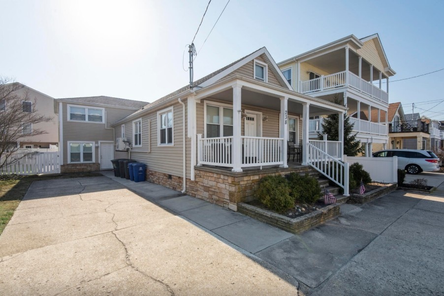 The exterior of a charming single-family home is showcased, featuring a welcoming porch with white railings. The driveway offers off-street parking, visible trash bins, and a landscaped area. Adjacent structures can be seen, indicating a residential neighborhood near the beach.