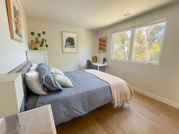 Dormitorio luminoso con grandes ventanales, luz natural, decoración moderna, suelos de madera, cama de matrimonio, ropa de cama acogedora, planta de interior y escritorio, ideal para estancias tranquilas y trabajo a distancia.
