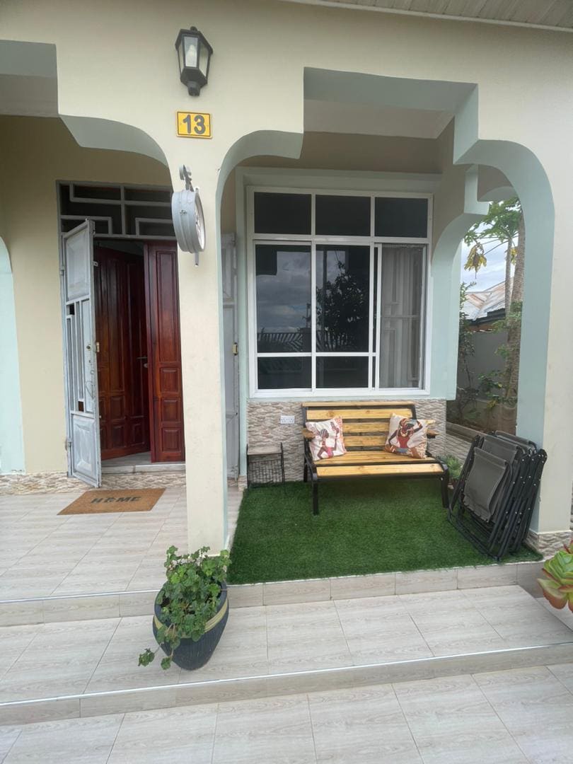 A welcoming entrance features a small bench adorned with decorative pillows. The area is complemented by a green patch of artificial grass and a potted plant. The entrance is framed by large windows, allowing natural light to illuminate the space.