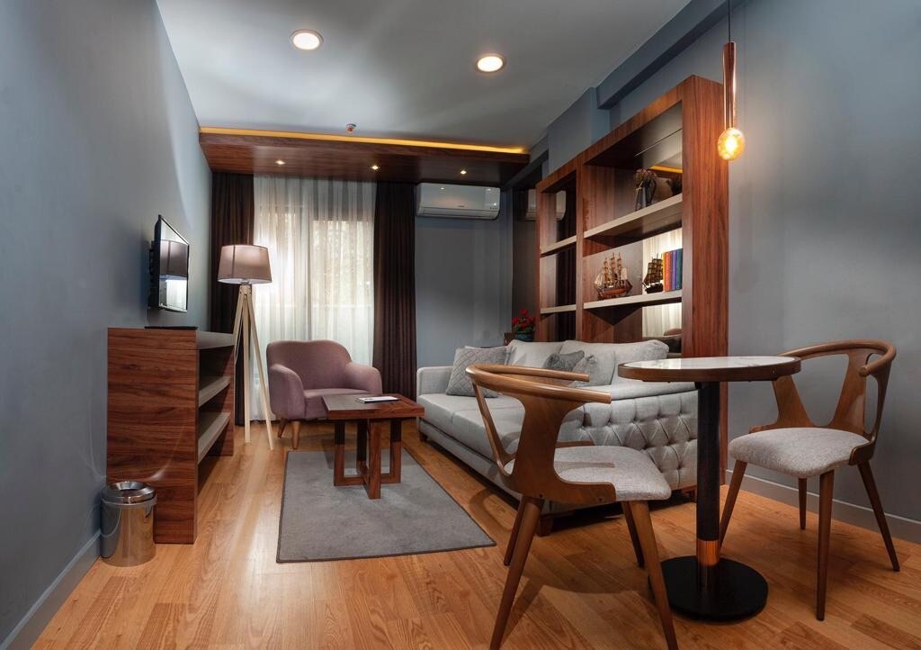 A stylish seating area features a light gray sofa and a small wooden coffee table positioned on a soft rug. Warm wooden shelves display decorative items. Natural light enters through sheer curtains, brightening the space equipped with ambient lighting and a wall-mounted television.