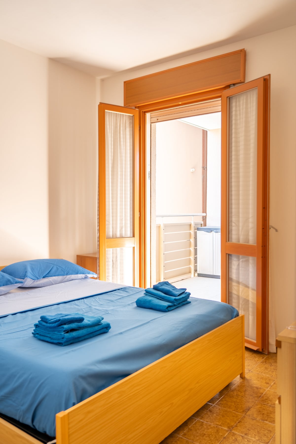 A bright bedroom features a spacious bed with blue bedding and neatly arranged towels on top. Natural light floods the room through large, open doors that lead to a small balcony. Light-colored walls provide a simple and welcoming backdrop.
