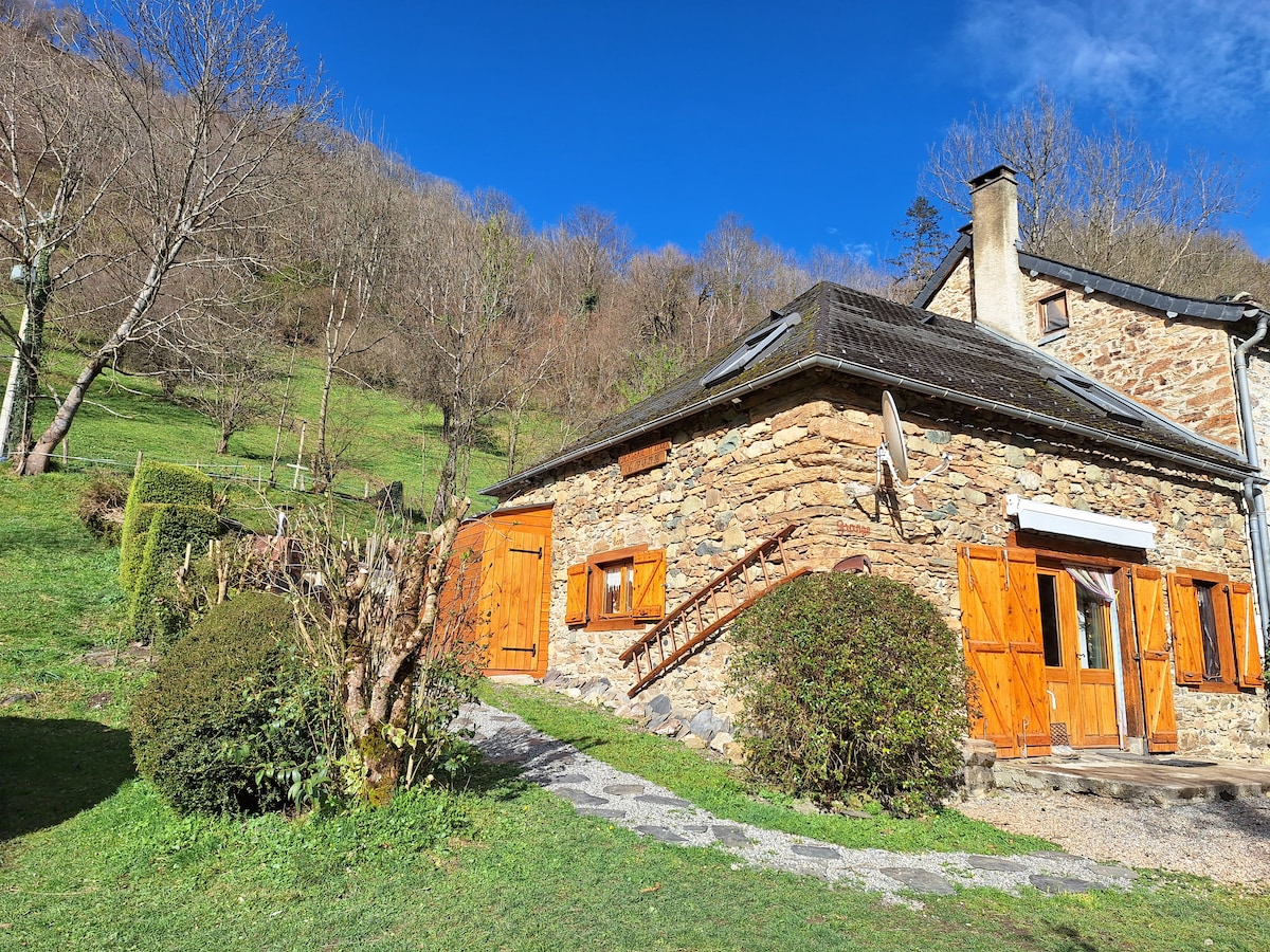 A rustic stone cottage is situated on a gently sloping green hillside, surrounded by bare trees. The exterior features wooden doors and windows, complemented by a winding pathway leading to the entrance. Natural light illuminates the structure against a clear blue sky.