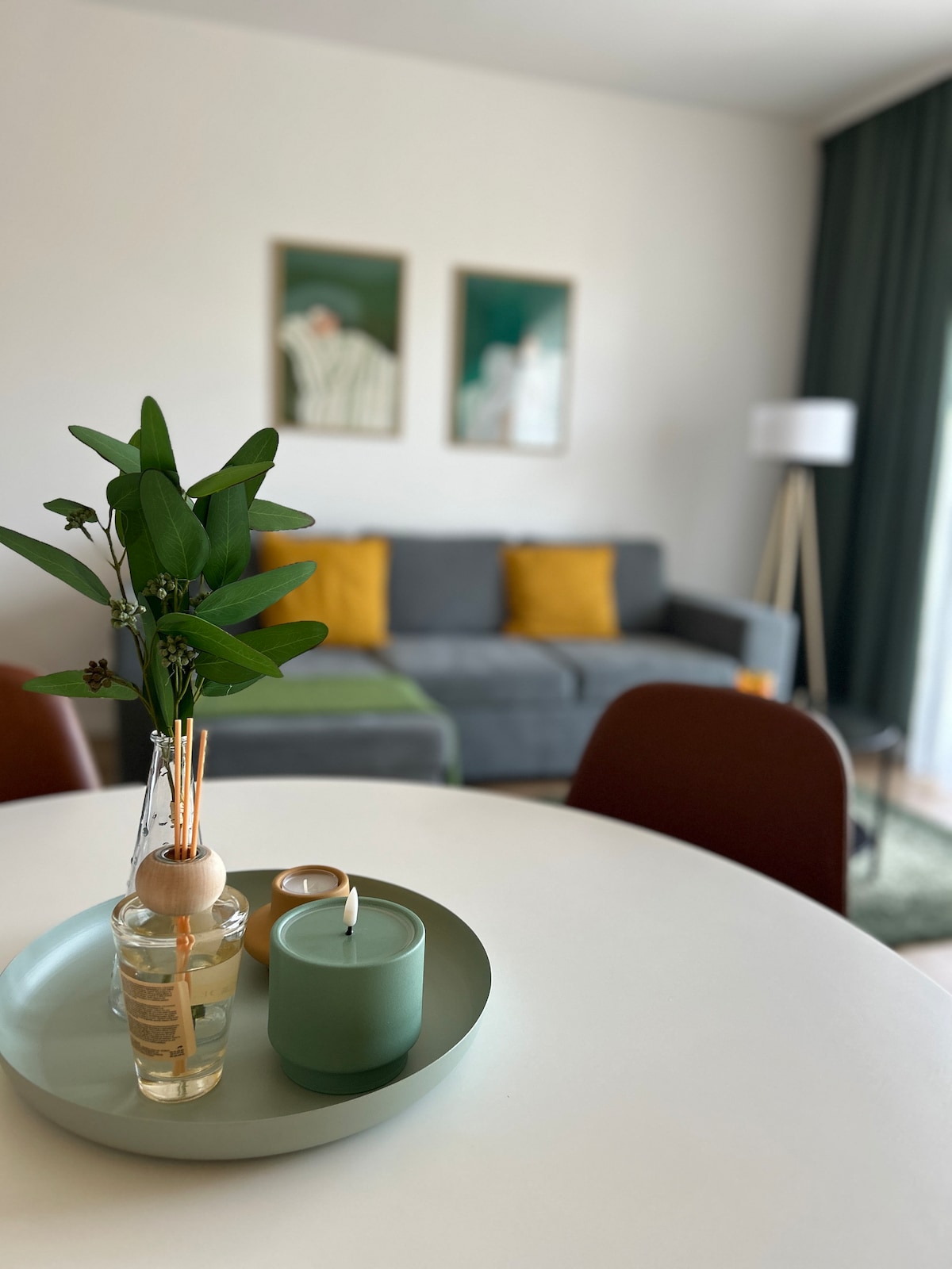 A modern living area is featured with a gray sofa adorned with yellow cushions. A round dining table is set with a green tray, showcasing a candle and a diffuser. Two art pieces are displayed on the wall, while natural light filters through a nearby green curtain.