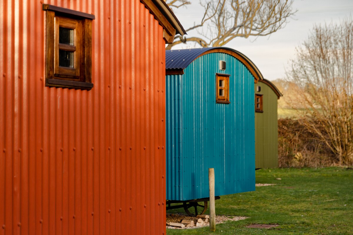 Three colorful shepherd's huts are displayed, featuring rounded roofs and corrugated metal exteriors in vibrant shades of orange, blue, and green. The huts are set in a grassy area with trees in the background, suggesting a serene rural environment.