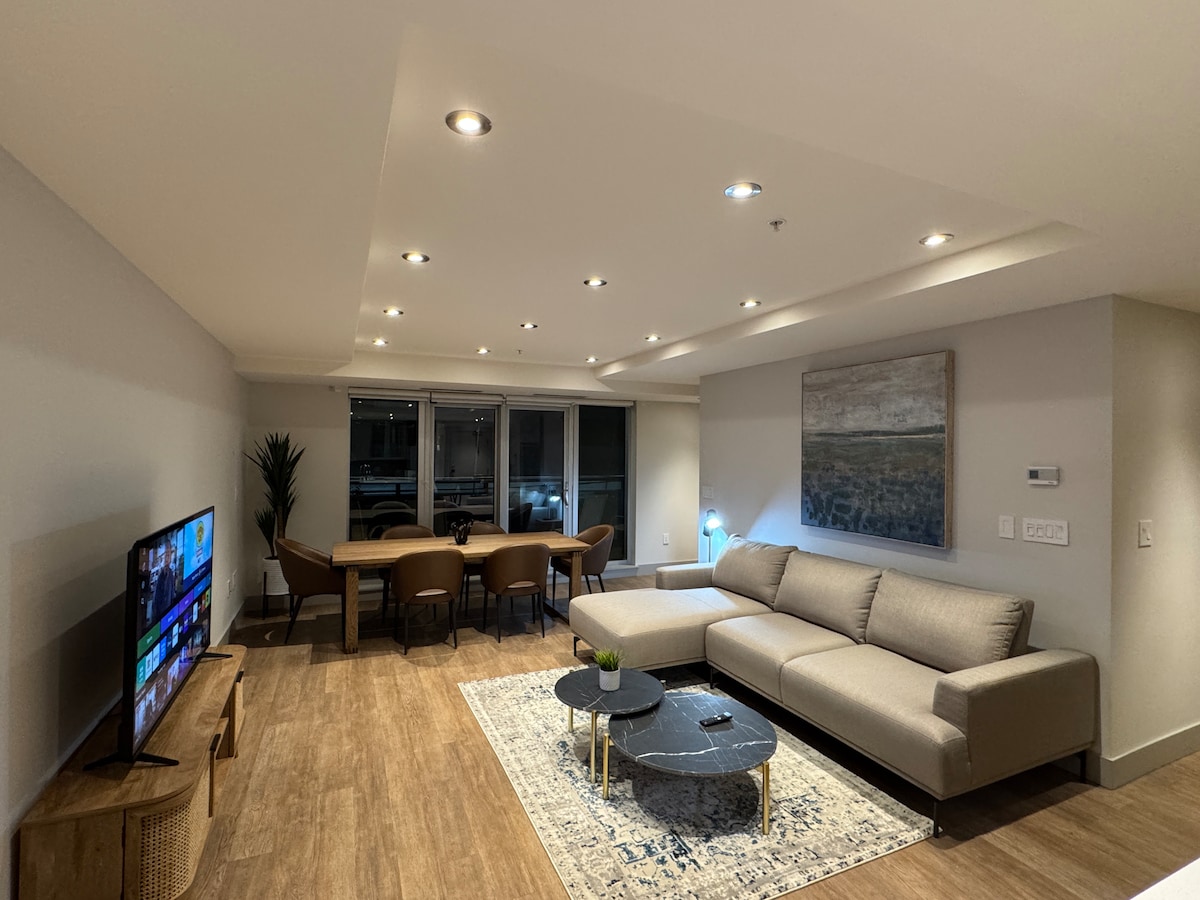 A modern living room is highlighted by a spacious beige sectional sofa, paired with a dark stone coffee table. A flat-screen TV is positioned on a light wood console. Soft lighting enhances the atmosphere, while large windows provide views of the outside.