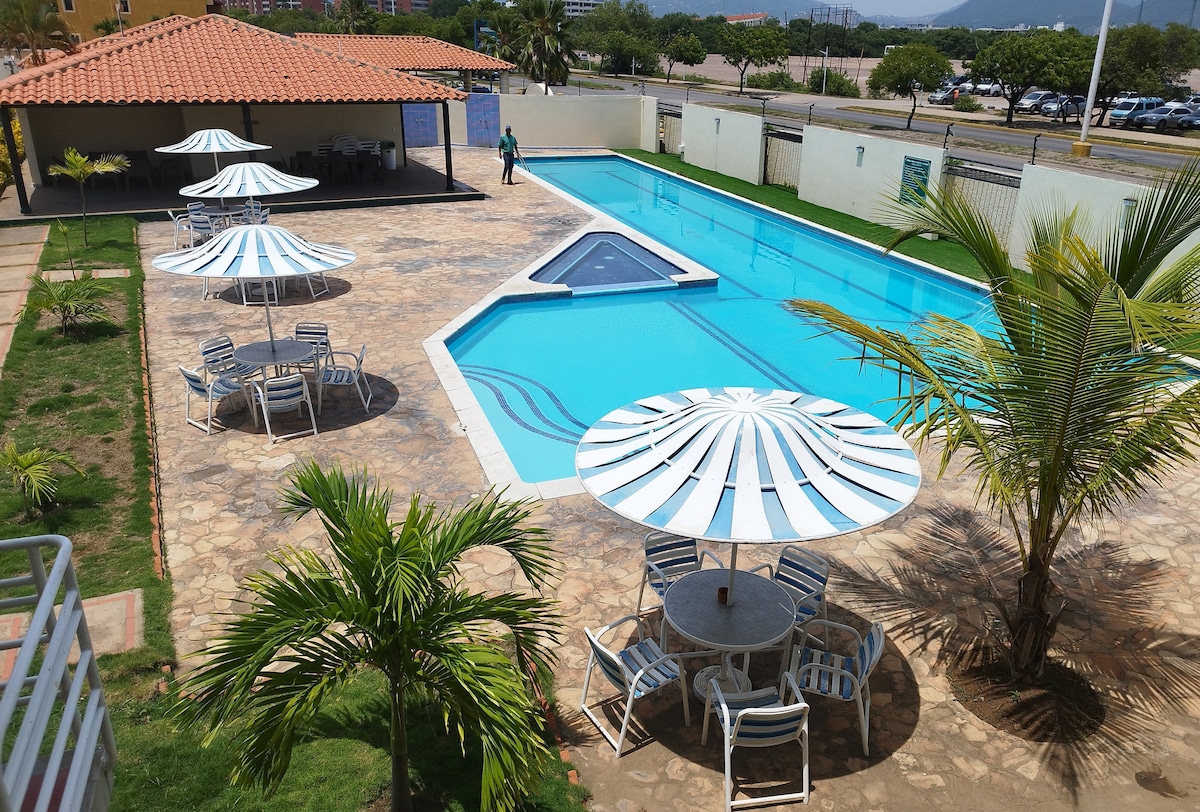 An inviting outdoor area features a large swimming pool with a unique triangular section. Several round tables and umbrellas are positioned on the poolside patio, surrounded by neatly maintained grass and palm trees. A shaded building is visible in the background.