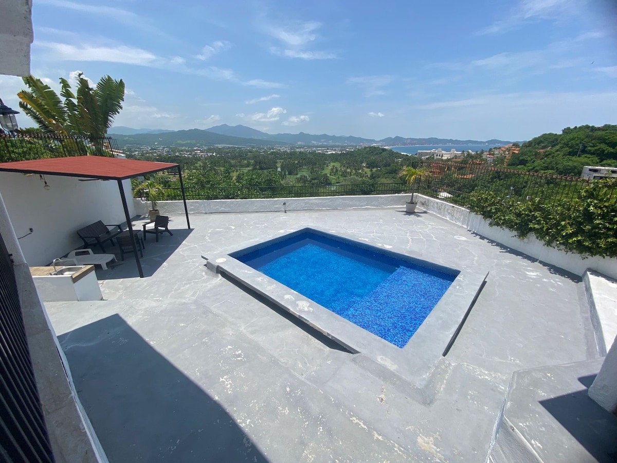 A private pool is set against a stunning panoramic backdrop, featuring clear blue water surrounded by a spacious patio. A shaded area with seating is visible nearby, enhancing the outdoor living space. Lush greenery and distant mountains complete the tranquil scene.