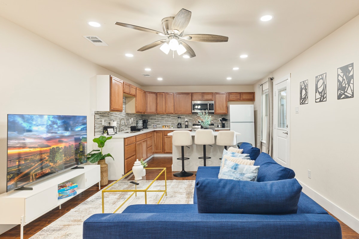 Moderný otvorený obytný priestor s plyšovými modrými sedadlami, elegantným stropným ventilátorom a veľkou inteligentnou televíziou. Plne vybavená kuchyňa zahŕňa raňajkový bar, spotrebiče z nehrdzavejúcej ocele a teplé drevené skrinky. Svetlé, čisté a ideálne na relax alebo zábavu.