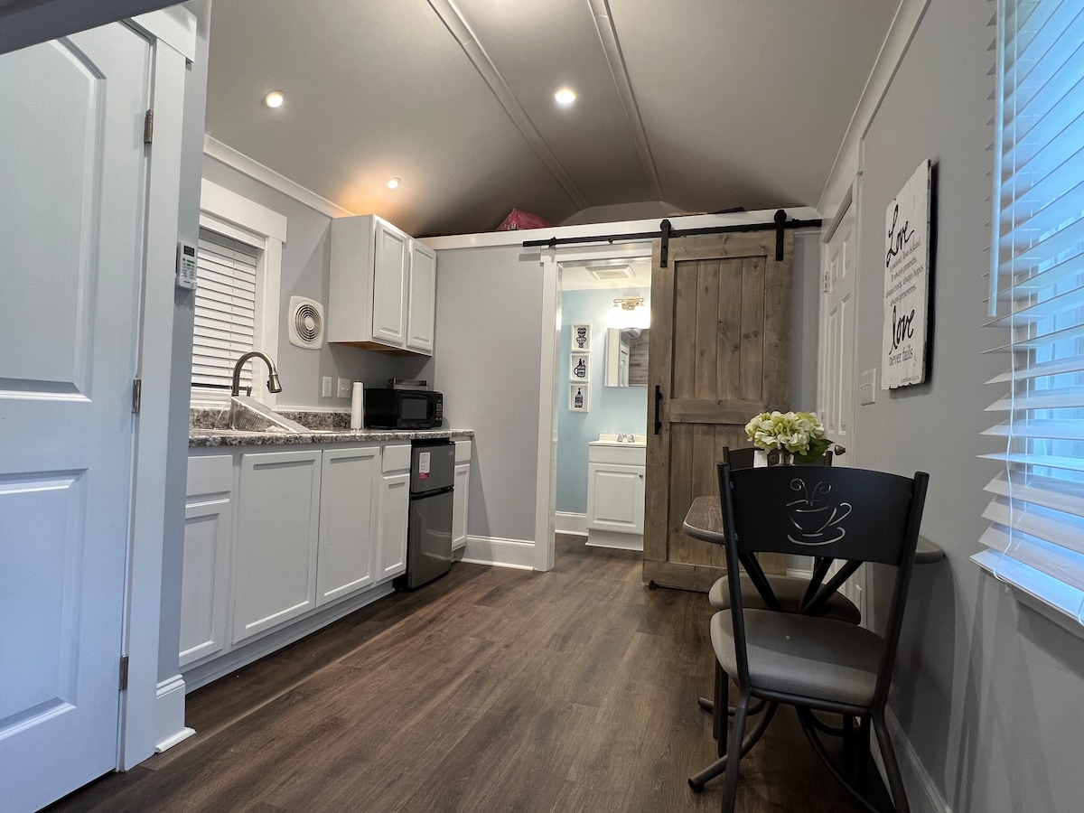 A modern kitchen area is visible with white cabinets and a granite countertop. A stainless steel refrigerator and a microwave are included. A sliding barn door leads to a bathroom. A small dining table with two chairs is positioned next to a window, allowing natural light in.