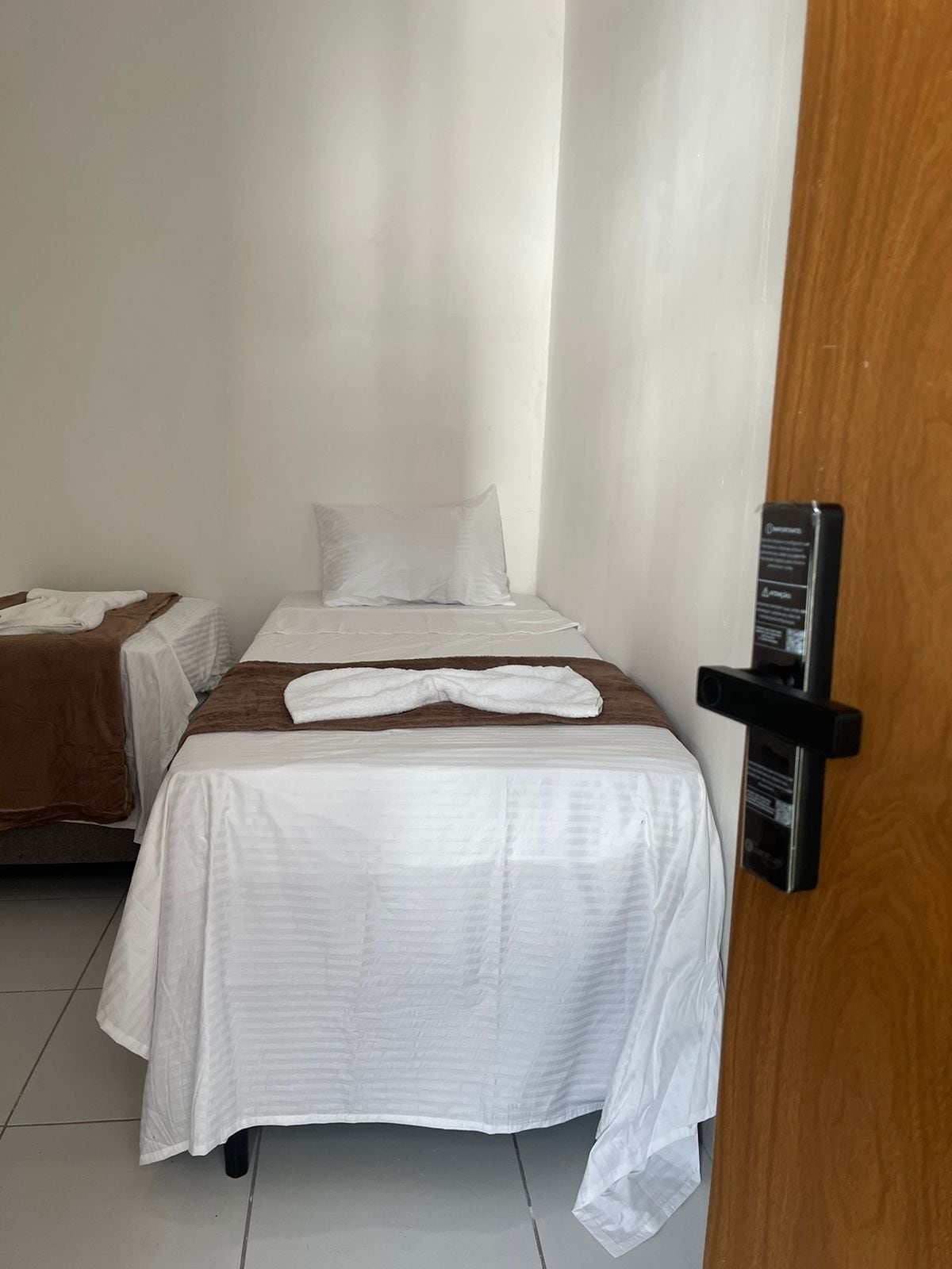 A modest room is presented, featuring three single beds, each covered with crisp white linens. Pillows are neatly arranged at the head of the beds. A door frame is visible to the right, leading to the rest of the accommodation. Natural light enters through a window, illuminating the space.