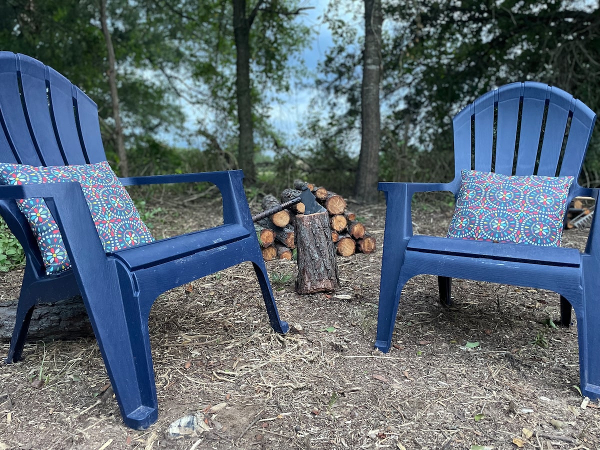 Two blue adirondack chairs are positioned around a small fire pit, with decorative cushions resting on the seats. A stacked pile of logs sits nearby, surrounded by a natural setting of trees and earthy ground.