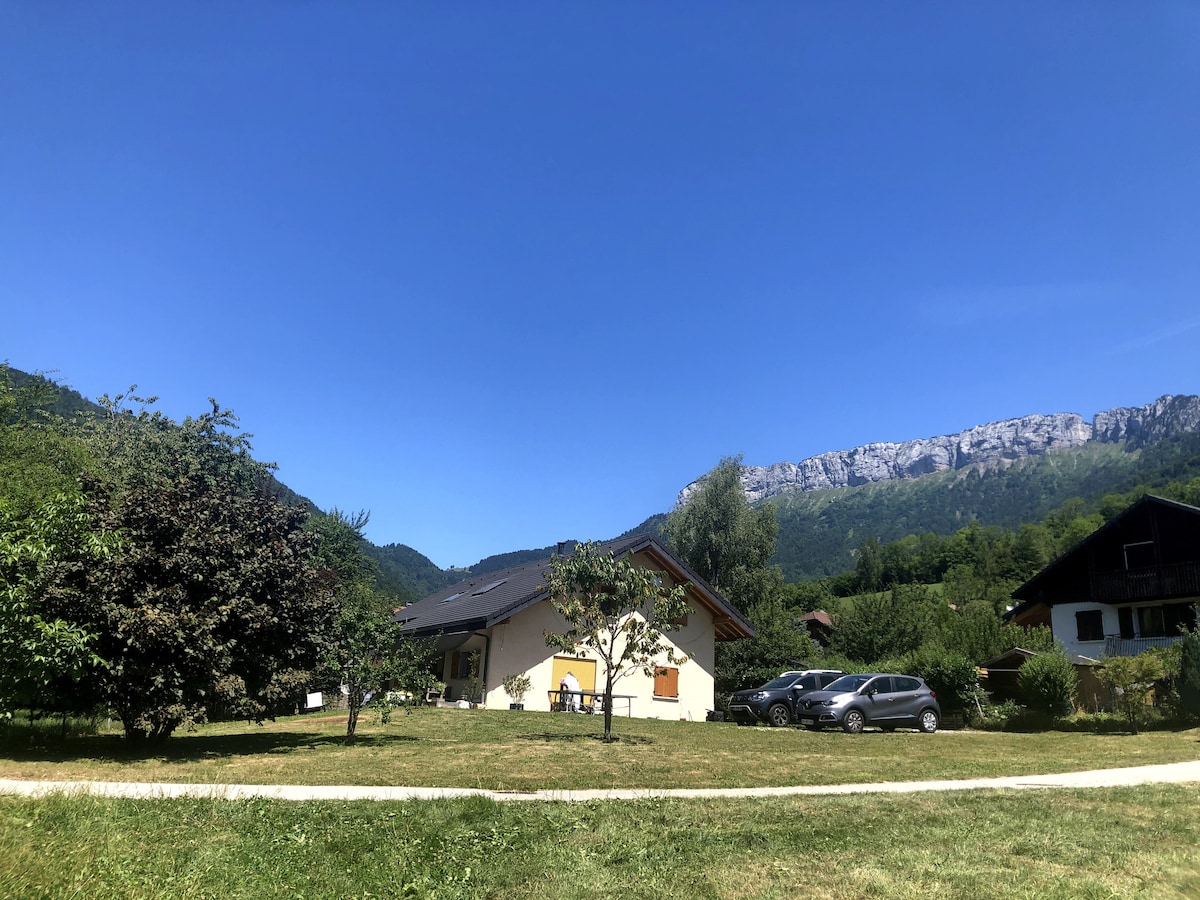 A bright yellow building with a sloped roof is surrounded by green grass and trees. The backdrop features mountains under a clear blue sky. Several parked cars can be seen adjacent to the structure, highlighting the spacious outdoor area.